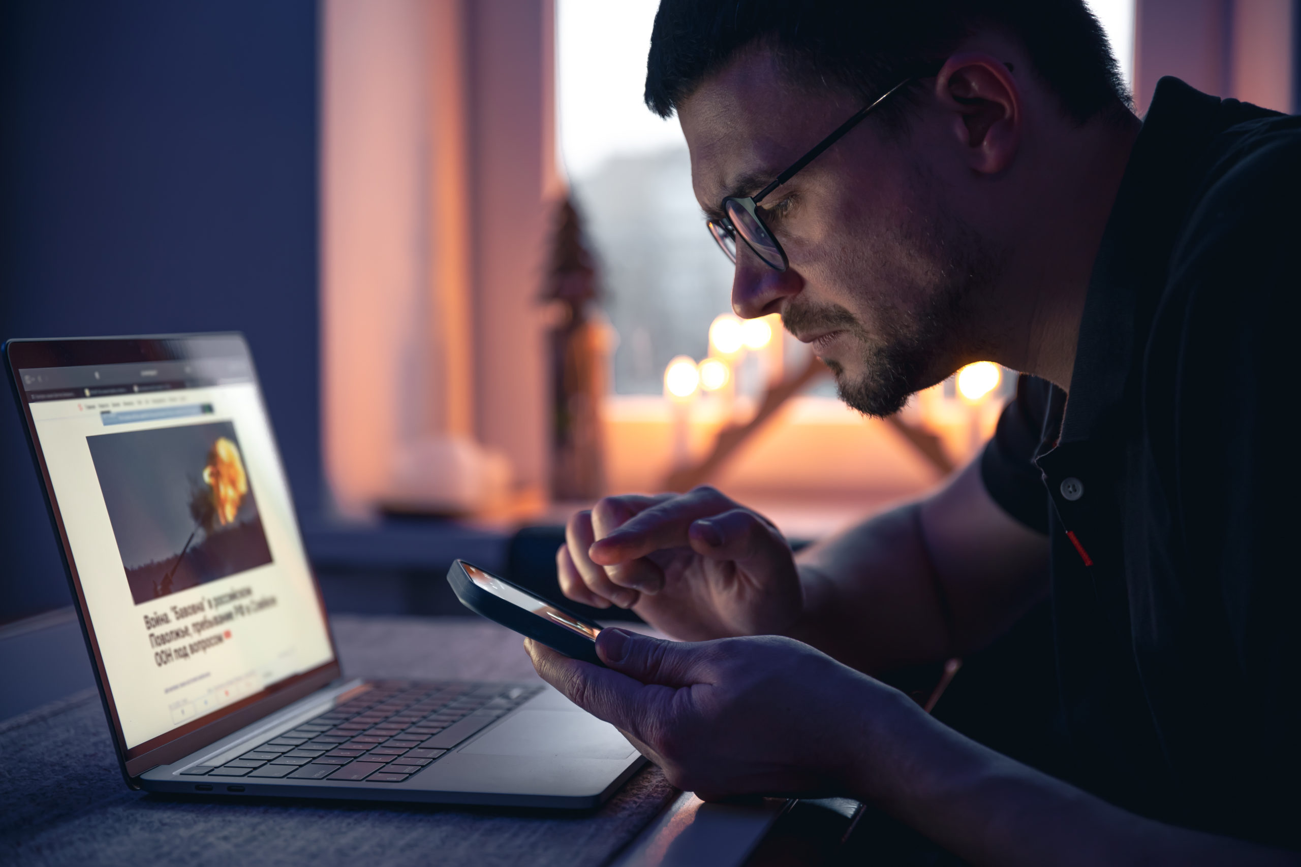 A man with a smartphone sits in front of a laptop late at night.